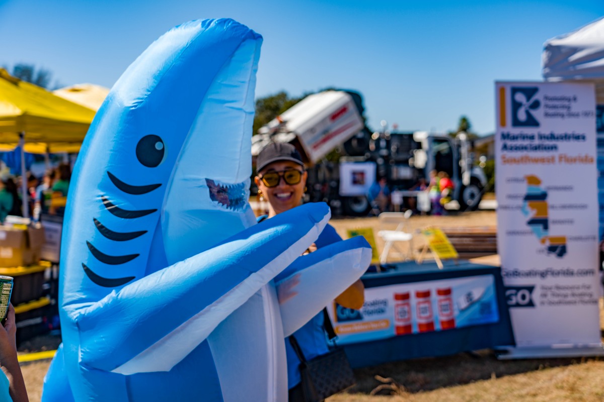 A smiling woman in an inflatable blue shark costume poses at the Waterways Festival in front of the Marine Industries Association of Southwest Florida booth.